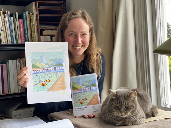 Wellington artist Hannah Webster holding the Our Wellington magazine and the original painting she did of Thorndon Pool for the cover. Her cat is asleep beside her on the desk.