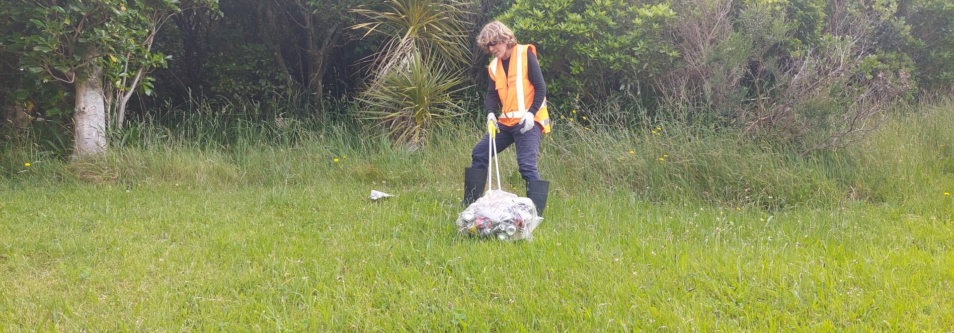 A man in high vis pulling a bag of rubbish across grass.