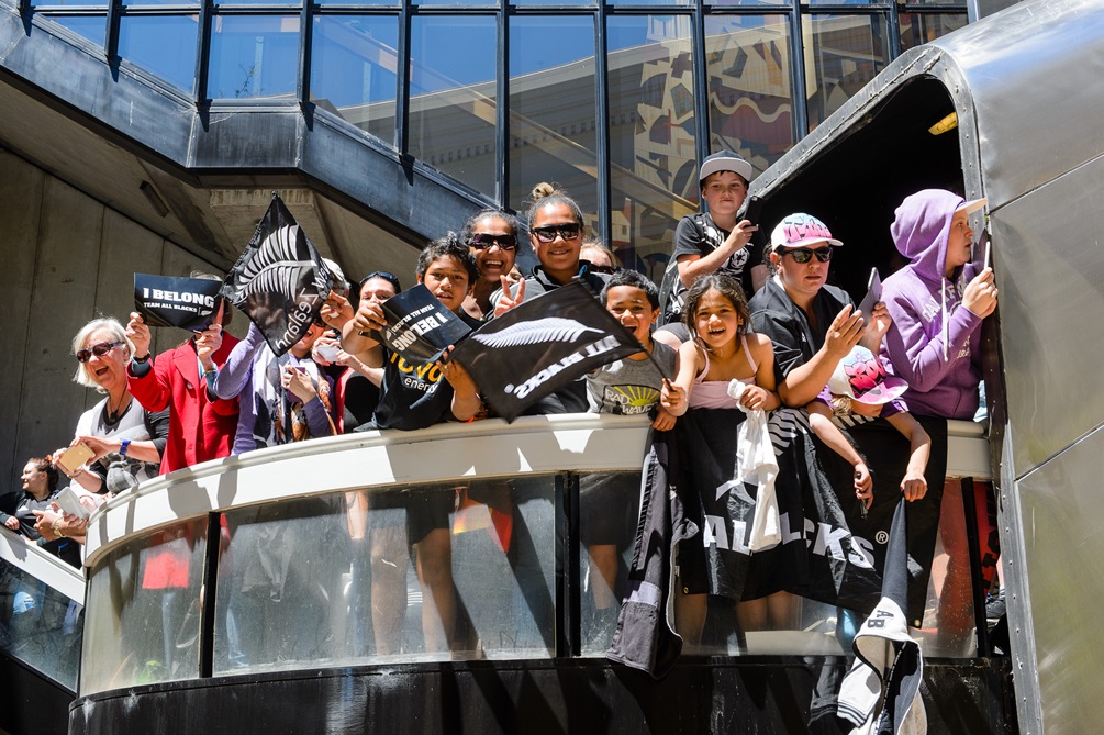 A crowd of rugby fans standing on a balcony wearing All Blacks merchandise and waving flags.