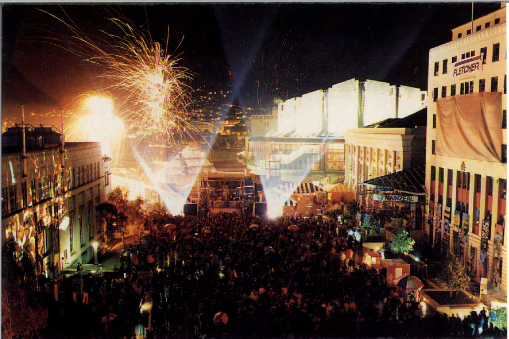 Crowds of people in Civic Square with bright lights and fireworks in the background.