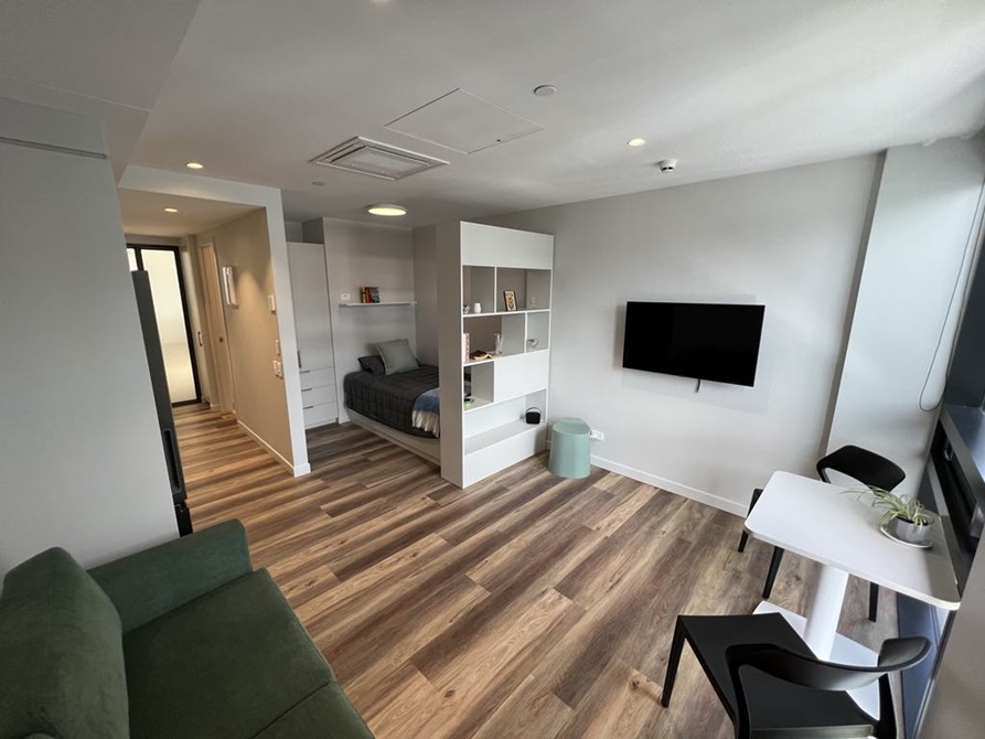 Studio apartment with a green couch in the foreground and a single bed with a tv and two person table.