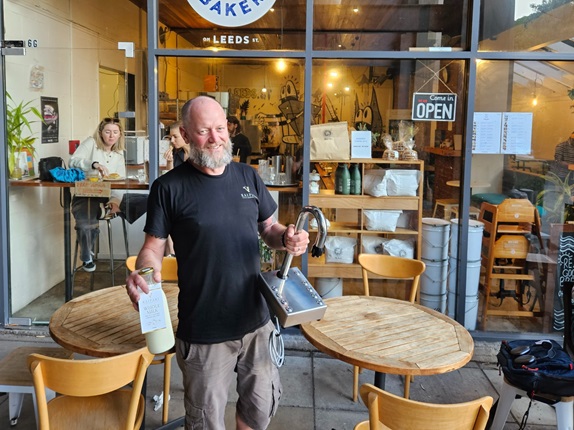 Man standing infront of a cafe holding a bottle of milk and a milk tap.
