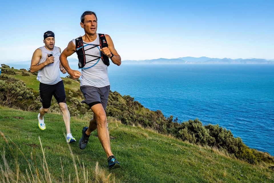 Two men running along ridge trail with views of ocean in the background.