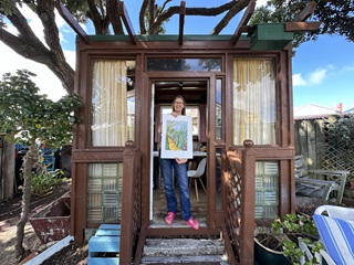 Wellington artist Carrie Carey, standing in the wooden doorframe of a tiny stand-alone room situated under a tall tree, holding her A3 screen-print of a vibrant yellow pathway surrounded by autumn coloured bush.