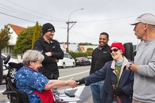 Two men who work for Downer construction stand happily onlooking as a woman in a wheelchair shakes hands with another woman. They are all in a suburban street setting.