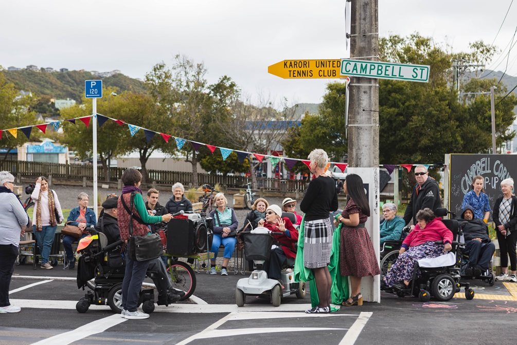 A large group of people, many in wheelchairs, gathered by the roadside with street decorations surrounding them.
