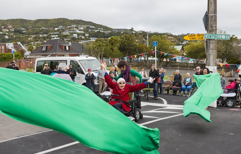 A very large green ribbon is cut by a woman wearing a read coat as she sits in a wheelchair on a pedestrian crossing. Onlookers stand on the roadside under street decorations.