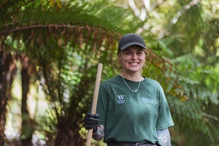 A woman in a green shirt smiling at the camera and holding the handle of a rake.