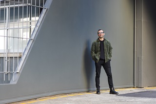 A man with glasses on, wearing a green overshirt and black tshirt, standing to the right of a grey wall.