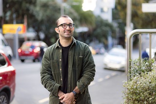 A man with glasses on, wearing a green overshirt and black tshirt, standing on a sidewalk with cars in the background.