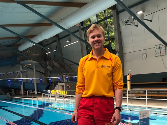A male lifeguard wearing his work uniform of a yellow polo long sleeve top and red shorts, standing infront of a swimming pool.