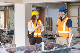 A woman talking to a man, both are wearing orange high vis jackets and hard hats.