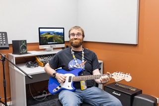 Image of Jamie sitting in a recording space with a blue electric guitar