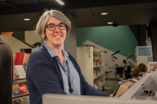 A woman wearing black glasses with short hair, smiling at the camera, surrounded by library books on shelves.