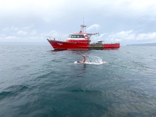 A person swimming in the water of the Foveaux Strait with a red boat in the background.