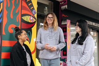 Three people standing on a street infront of a brightly coloured building painted with a mural. 