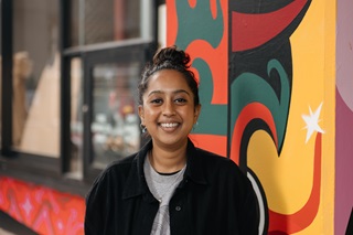 A woman standing infront of a brightly painted building, smiling at the camera. She is wearing a black jacket over the top of a grey sweatshirt with her hair pulled into a bun.