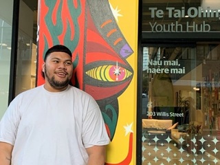 A man standing infront of a brightly painted building, smiling to the side of the camera. He is wearing a white tshirt and has dark features.