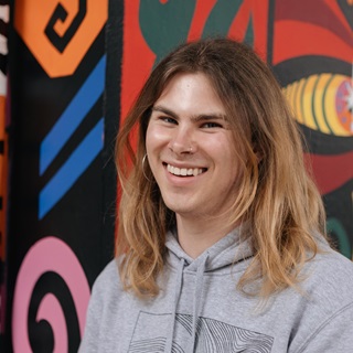 A person standing infront of a brightly painted building, smiling directly at the camera. They are wearing a grey hoodie with shoulder length brown and blonde hair.