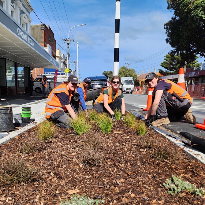 Group of people in high vis kneeling on the side of the road, tending to a garden.