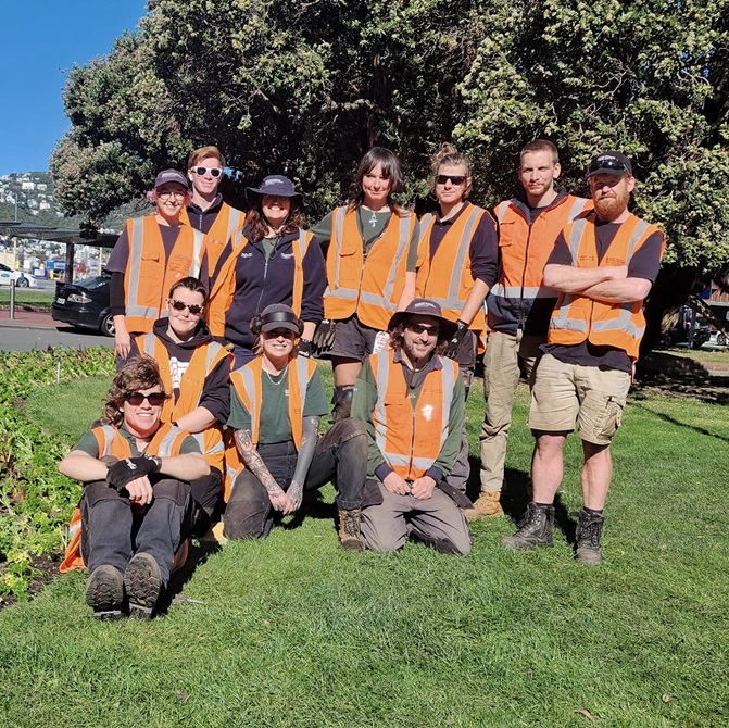 A group of nine people wearing high vis vests and gardening uniforms, standing on grass.