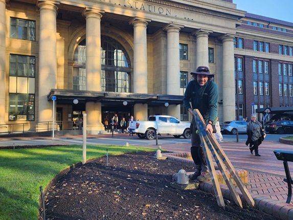 Gardener Tom Box wearing all green and a hat, tending to a garden outside of the railway station.