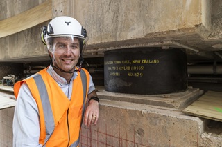 Man wearing a white collared shirt, orange high vis vest and white hard hat standing infront of a base isolator.