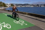 Person cycling on top of a green bike lane next to the harbour with Wellington city in the distance.