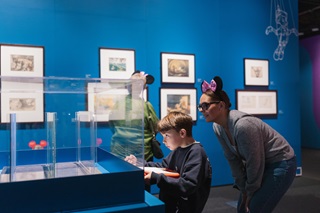 Child sketches model at Disney exhibition with adult watching while wearing Minnie Mouse ears