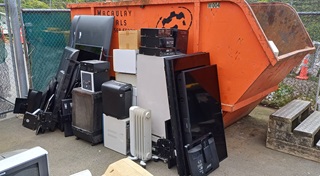 Old electronics from heaters to tvs, leaning against an orange skip bin outdoors.