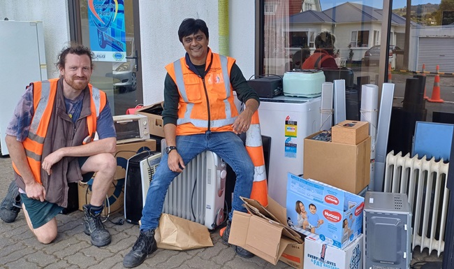 Two men wearing high vis sitting on top of a pile of ewaste on the sidewalk.