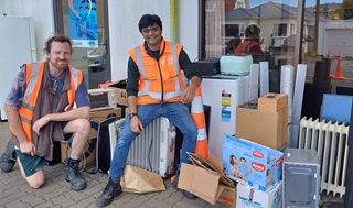 Two men wearing high vis sitting on top of a pile of ewaste on the sidewalk.