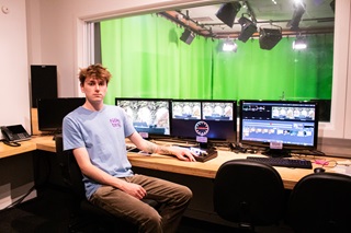 A young man in a blue tshirt, sitting on a desk chair infront of a row of tv screens. In the background, a green screen has been set up in a studio.