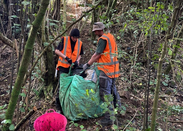 Two people wearing high vis vests and gardening clothes, standing inside of a forest. One is holding a green bag open while the other inserts rubbish into it.