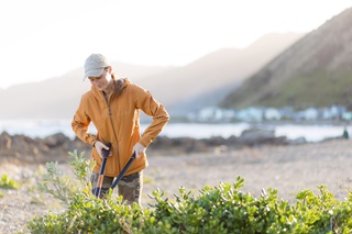 A woman in an orange jacket, chopping back plants on the coast.