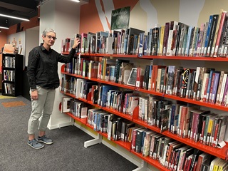 A woman wearing a black top and beige pants standing infront of a bookshelf.