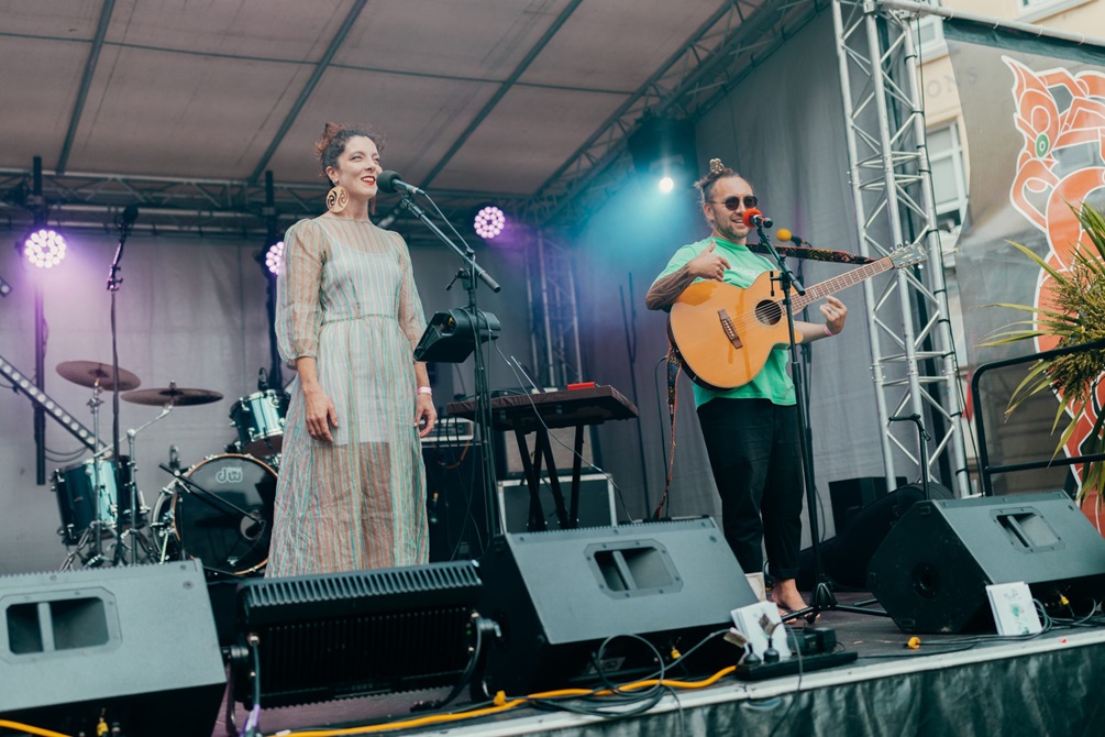 Two performers on stage, one is holding a guitar and the other is singing.