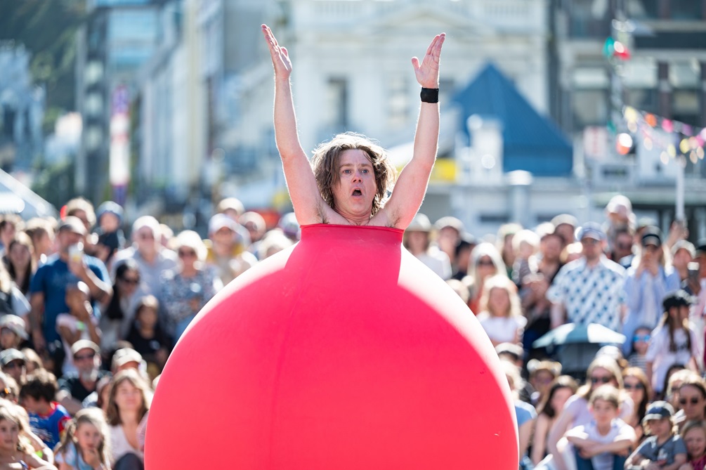 A person dressed in a giant red bauble with a crowd of people watching them perform.