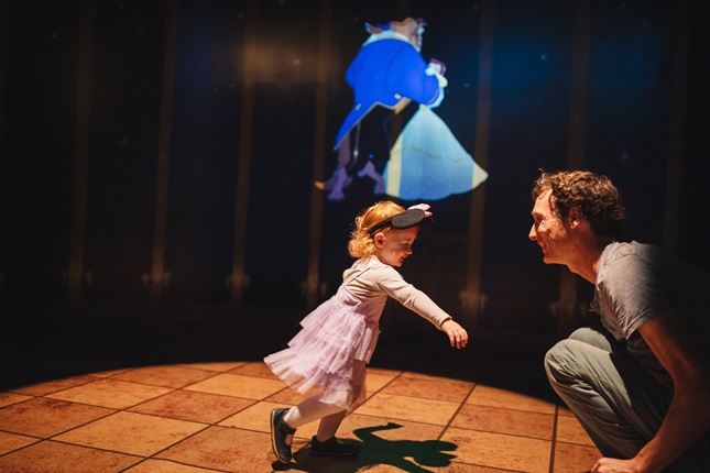 Little girl wearing a pink dress running towards a man in a lit up area within an exhibition.