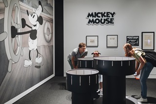 Three people looking into a Zoetrope at the Disney Exhibition with Steamboat Willie on the wall.