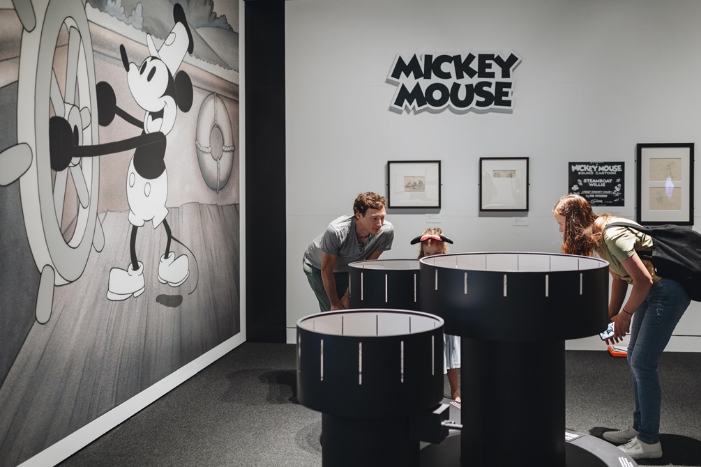 Three people looking into a Zoetrope at the Disney Exhibition with Steamboat Willie on the wall.