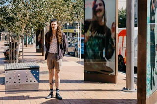 A casually dressed person stands on a sunny pedestrian street with trees and benches, next to a reflective glass panel showing their image.
