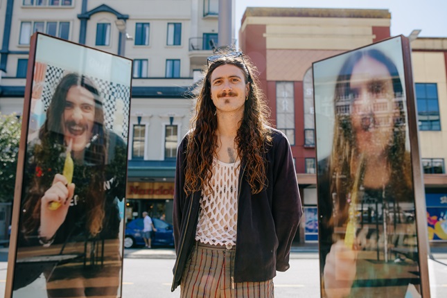 A person with long hair stands between two reflective glass panels displaying their mirrored images, with buildings in the background.
