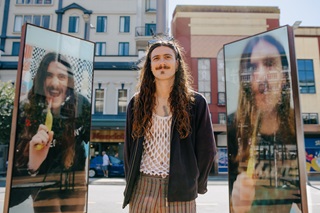 A person with long hair stands between two reflective glass panels displaying their mirrored images, with buildings in the background.