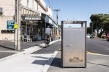Rubbish bin in the middle of an urban street with shops in the background and a person riding their bike.