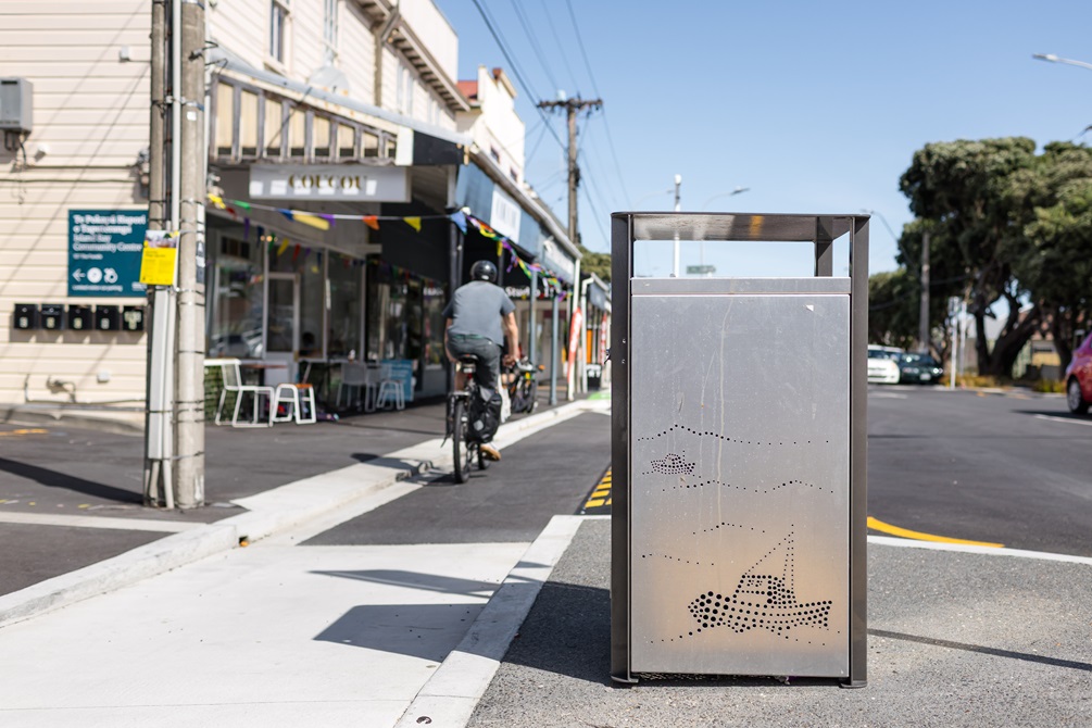 Rubbish bin in the middle of an urban street with shops in the background and a person riding their bike.