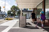 Rubbish bin in the middle of an urban street with two people on the right hand side walking past.