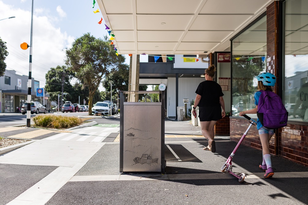 Rubbish bin in the middle of an urban street with two people on the right hand side walking past.
