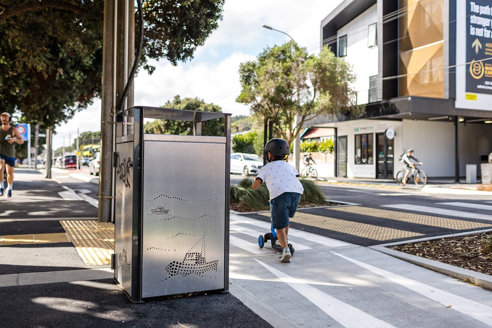 Rubbish bin in the middle of an urban street with a small child riding a scooter next to it.
