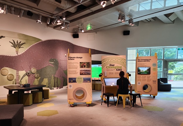 Person sitting at an exhibition inside a museum with chairs and a table to one side.
